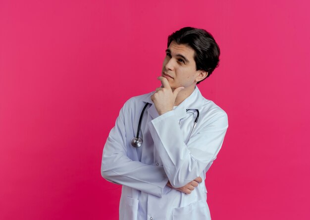 복사 공간이 분홍색 벽에 고립 된 턱을 만지고 측면을보고 의료 가운과 청진기를 입고 사려 깊은 젊은 남성 의사