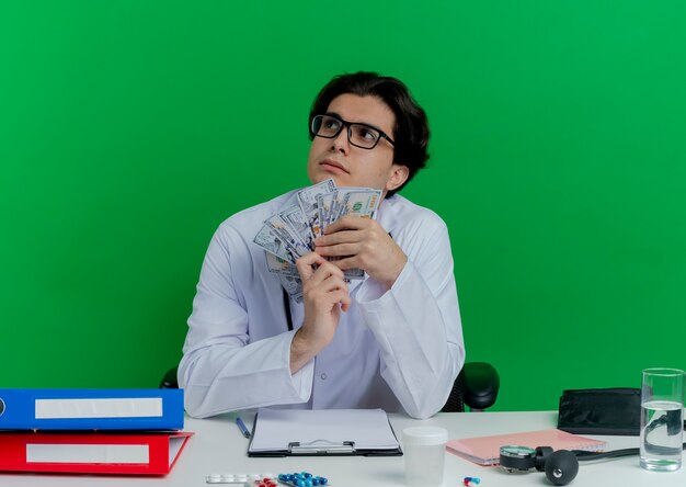 Thoughtful young male doctor wearing medical robe and stethoscope with glasses sitting at desk with medical tools holding money looking at side isolated on green wall