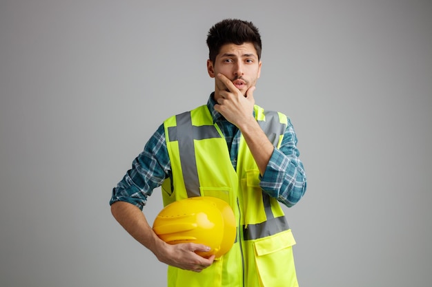 Thoughtful young male engineer wearing uniform holding safety helmet looking at camera while keeping hand on chin isolated on white background