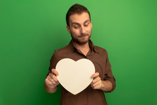 Free photo thoughtful young man holding and looking at heart shape isolated on green wall