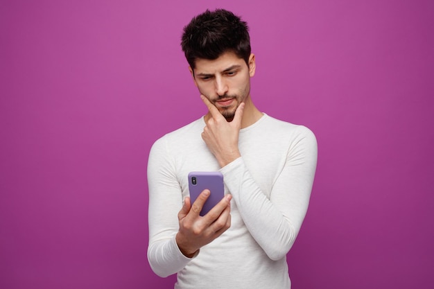 Вдумчивый молодой человек держит и смотрит на мобильный телефон, держа руку на подбородке, изолированный на фиолетовом фоне