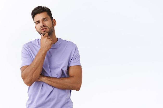 Thoughtful young man picking something in store, touching chin pensive squinting, considering what choice best, making opinion, express judgement, picking variant, pondering over white wall