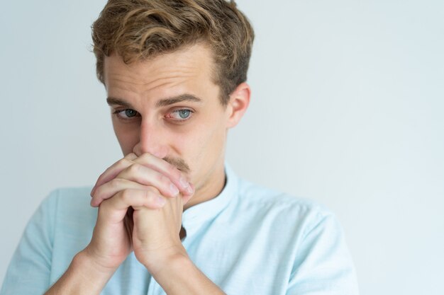 Thoughtful young man touching mouth with clenched hands and looking away.