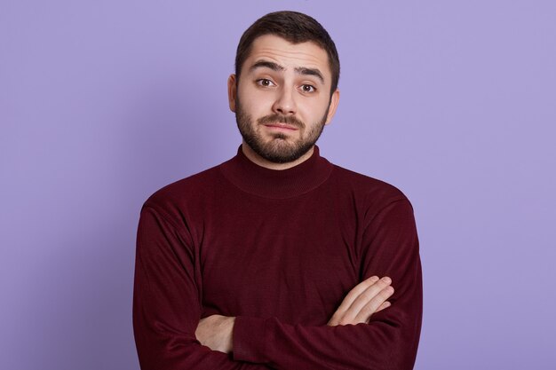 Thoughtful young man with skeptic, doubtful, distrustful look posing against lilac background with folded hands, wearing burgundy sweater