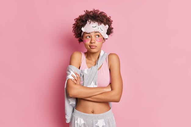 Thoughtful young pretty Afro American woman keeps arms folded has deep thoughts about something applies collagen patches under eyes