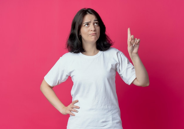 Thoughtful young pretty caucasian girl looking and pointing up and keeping another hand on waist isolated on crimson background