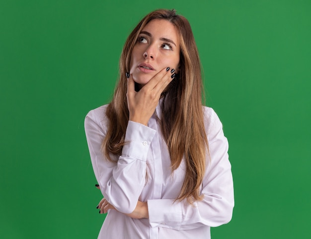 Thoughtful young pretty caucasian girl puts hand on chin and looks at side on green 