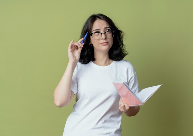 Thoughtful young pretty caucasian girl wearing glasses looking at side touching head with pen and holding note pad isolated on olive green background with copy space