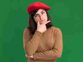 Free photo thoughtful young pretty caucasian girl with beret hat holding chin and looking at camera on green