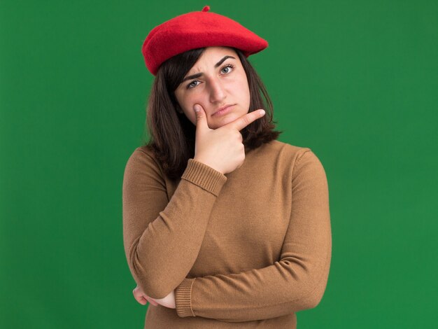 Thoughtful young pretty caucasian girl with beret hat holding chin and looking at camera on green
