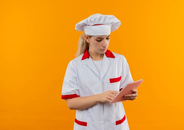 オレンジ色のスペースに分離されたメモ帳に指を置いて見ているシェフの制服を着た思いやりのある若いかわいい料理人