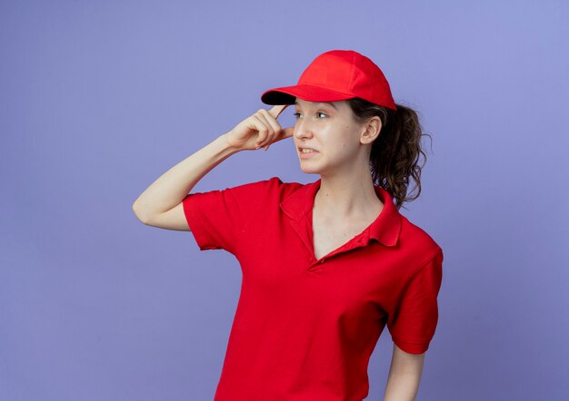 Thoughtful young pretty delivery girl wearing red uniform and cap putting finger on head looking at side isolated on purple background with copy space