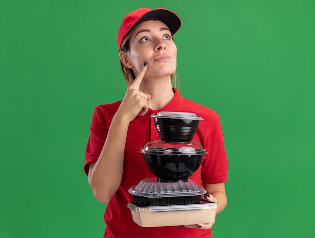 Thoughtful young pretty delivery woman in uniform holds food containers on food package looking up isolated on green wall