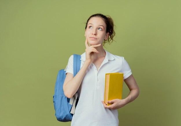 Thoughtful young pretty female student wearing back bag holding book looking up and touching chin isolated on green background with copy space