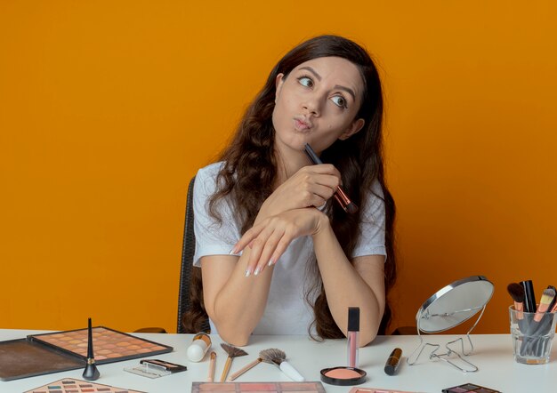 Thoughtful young pretty girl sitting at makeup table with makeup tools holding blush brush and touching chin with it looking at side isolated on orange background