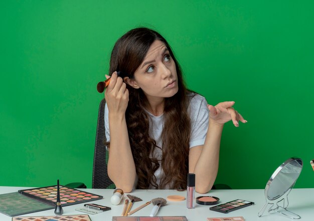 Thoughtful young pretty girl sitting at makeup table with makeup tools touching head with blush brush looking at side and keeping hand in air isolated on green background