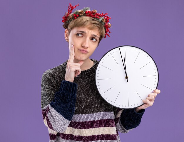 Thoughtful young pretty girl wearing christmas head wreath holding clock keeping hand on chin looking at side isolated on purple background