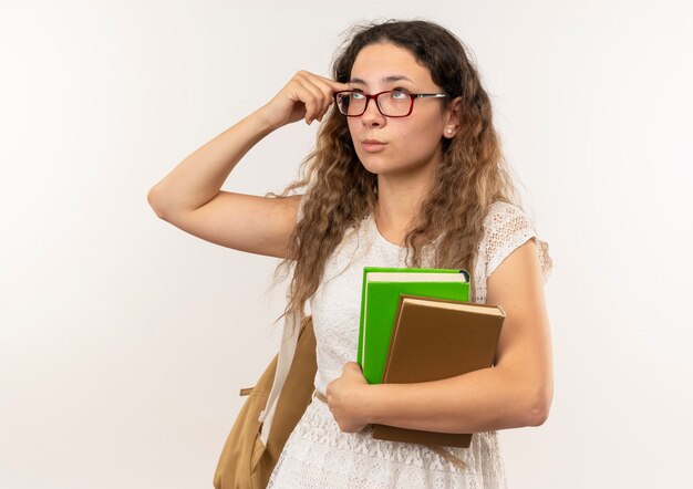 Thoughtful young pretty schoolgirl wearing glasses and back bag holding books looking up putting finger on temple isolated on white wall
