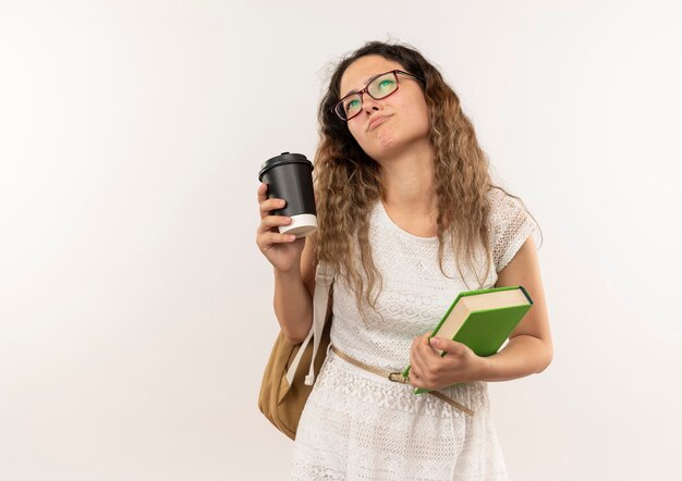 Thoughtful young pretty schoolgirl wearing glasses and back bag looking up holding coffee cup and book isolated on white wall