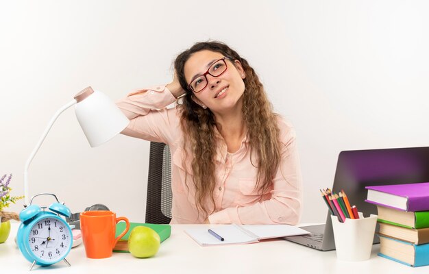 Thoughtful young pretty schoolgirl wearing glasses sitting at desk with school tools doing her homework putting hand on head looking at side isolated on white wall