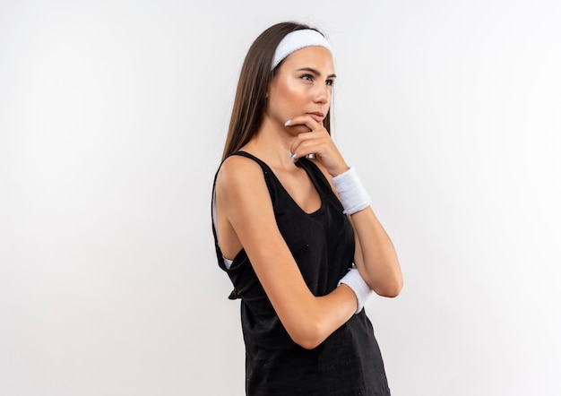 Thoughtful young pretty sporty girl wearing headband and wristband putting hand on chin looking at side on white space 