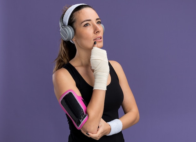 Free photo thoughtful young pretty sporty girl wearing headband wristbands headphones and phone armband with injured wrist wrapped with bandage looking up touching chin