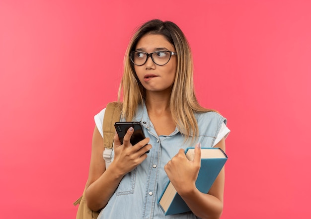 Thoughtful young pretty student girl wearing glasses and back bag holding mobile phone and book looking at side and biting lip isolated on pink wall