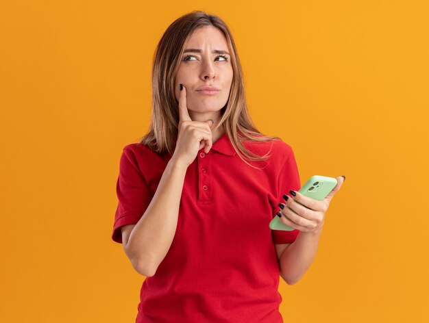 Thoughtful young pretty woman puts finger on chin and holds phone isolated on orange wall