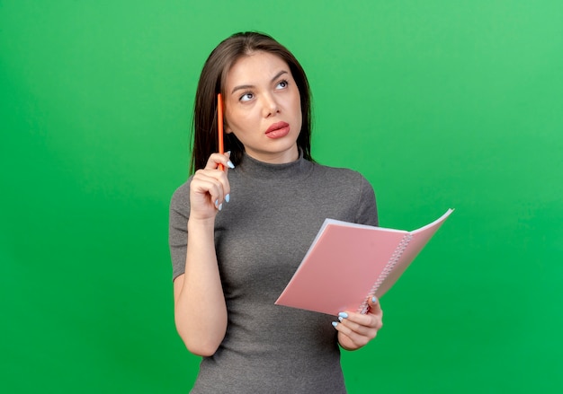 Thoughtful young pretty woman touching head with pen and holding open note pad looking up isolated on green background with copy space