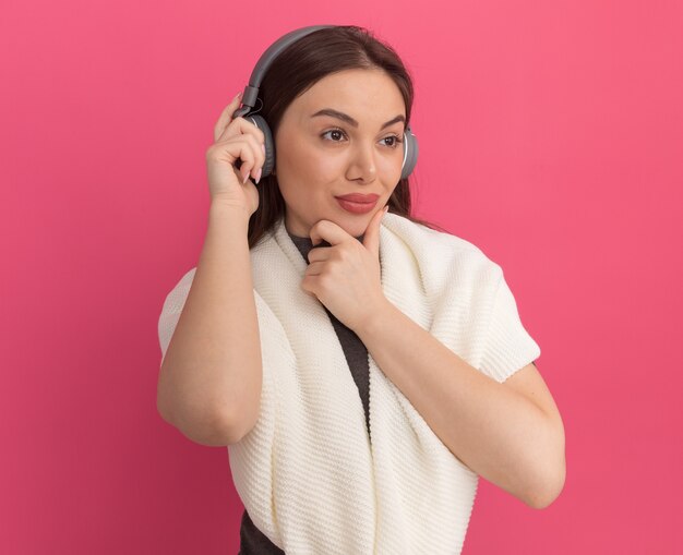 Thoughtful young pretty woman wearing and grabbing headphones looking at side keeping hand on chin 