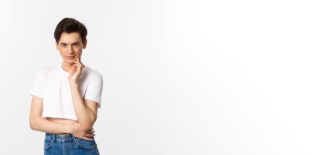 Thoughtful young queer man wearing crop top smiling and looking at camera cunning having an idea standing over white background