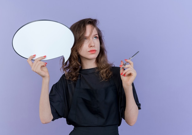 Thoughtful young slavic female barber wearing uniform holding chat bubble and scissors looking at side isolated on purple background with copy space