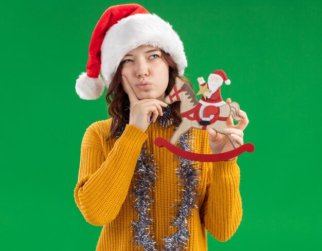 Thoughtful young slavic girl with santa hat and with garland around neck puts finger on chin and holds santa on rocking horse decoration looking at side isolated on green background with copy space