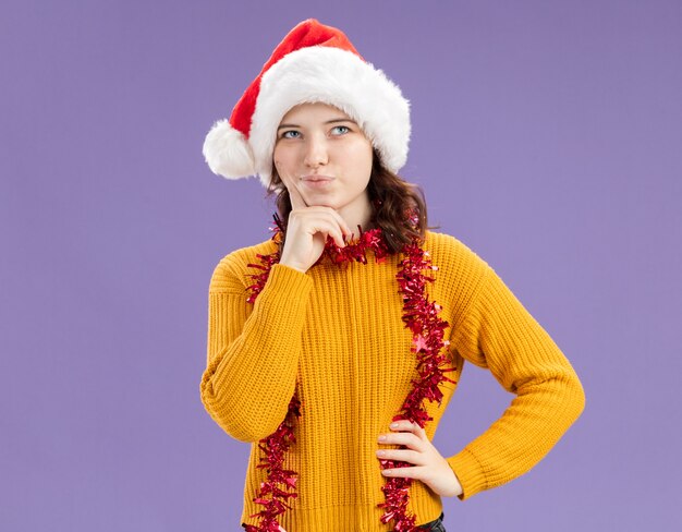 Thoughtful young slavic girl with santa hat and with garland around neck puts finger on chin and looks up isolated on purple wall with copy space