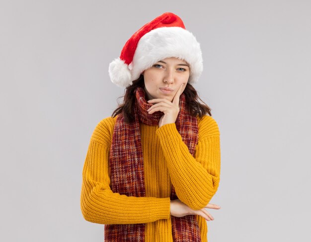 thoughtful young slavic girl with santa hat and with scarf around neck holds chin and looks at camera isolated on white background with copy space