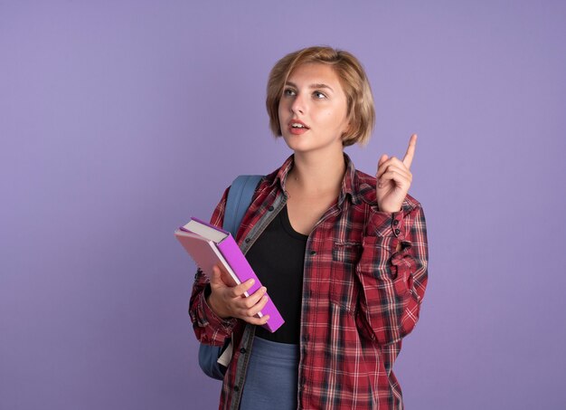 Thoughtful young slavic student girl wearing backpack holds book and notebook points up looking at side 