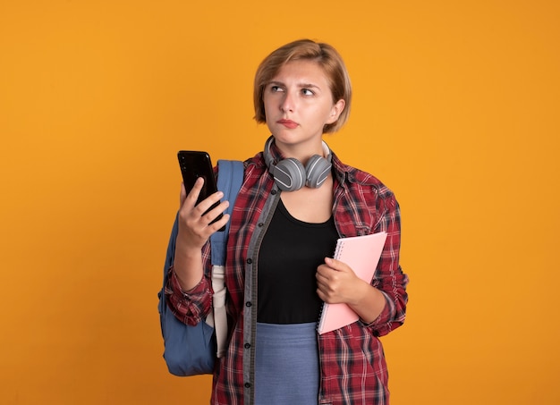 バックパックを着たヘッドフォンを付けた思慮深い若いスラブ学生の女の子は、ノートと携帯電話を横に見ている