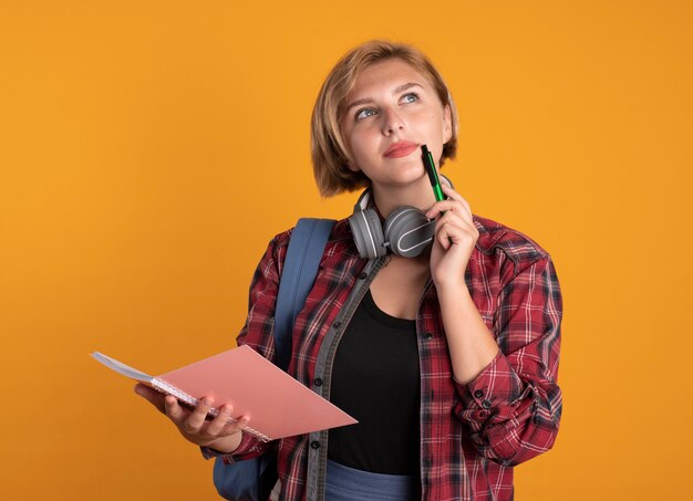 Thoughtful young slavic student girl with headphones wearing backpack holds pen and notebook 