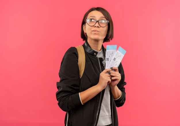 Задумчивая молодая студентка в очках и задней сумке с билетами на самолет смотрит в сторону, изолированную на розовой стене