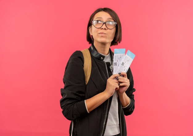 Thoughtful young student girl wearing glasses and back bag holding airplane tickets looking at side isolated on pink wall