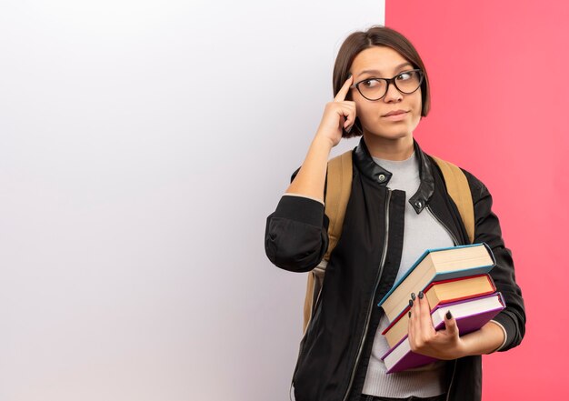 Задумчивая молодая студентка в очках и задней сумке держит книги, стоящие перед белой стеной, глядя в сторону пальцем на виске, изолированном на розовой стене
