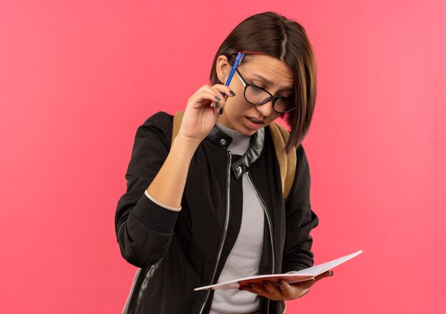 Thoughtful young student girl wearing glasses and back bag holding and looking at note pad touching head with pen isolated on pink wall