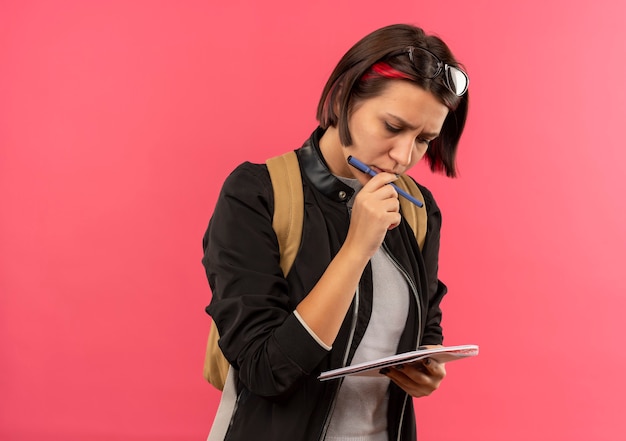 Thoughtful young student girl wearing glasses on head and back bag holding pen and note pad looking at note pad touching her lips with finger isolated on pink wall