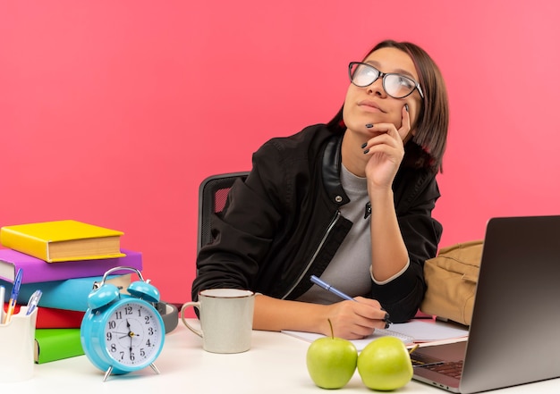 Thoughtful young student girl wearing glasses sitting at desk with university tools holding pen looking up putting hand on chin doing homework isolated on pink wall