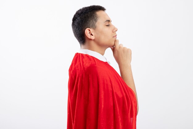 Thoughtful young superhero boy in red cape standing in profile view touching chin looking straight isolated on white background with copy space