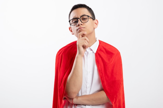 Thoughtful young superhero boy in red cape wearing glasses looking at camera keeping hand on chin isolated on white background with copy space