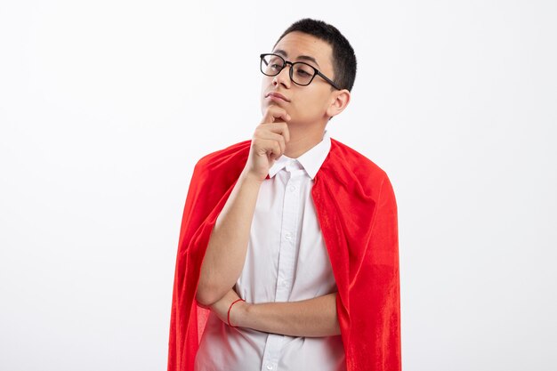 Thoughtful young superhero boy in red cape wearing glasses looking at side touching chin isolated on white background with copy space