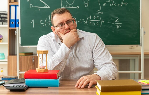 вдумчивый молодой учитель в очках сидит за столом со школьными принадлежностями в классе, держа руку за подбородок и на столе, глядя в сторону