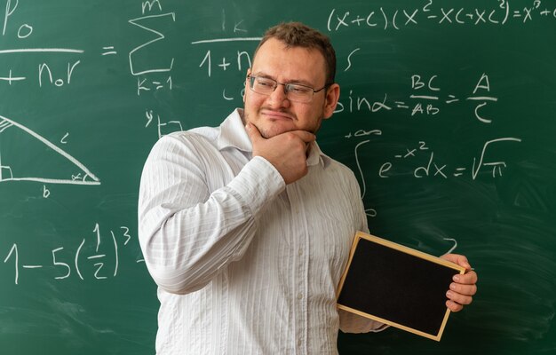 thoughtful young teacher wearing glasses standing in front of chalkboard in classroom holding mini blackboard keeping hand on chin looking at side
