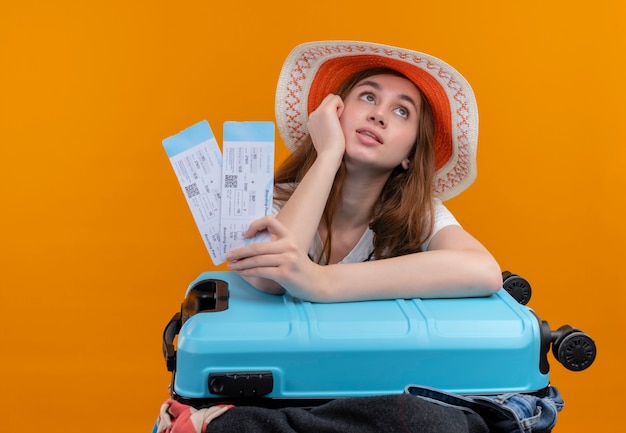 Thoughtful young traveler girl wearing hat holding airplane tickets and putting arm on suitcase on isolated orange wall